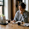Woman tasting coffee and writing in journal at kitchen table